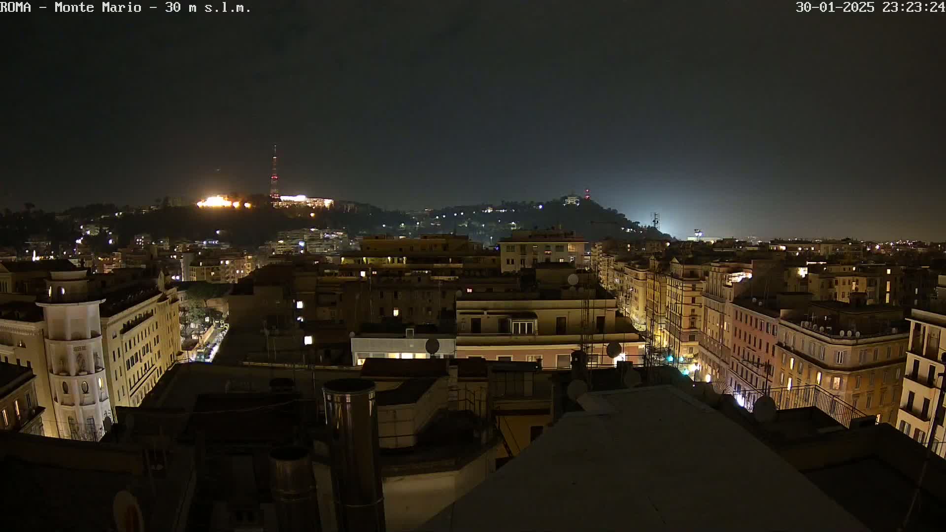 A nighttime view of a city, with dimly lit buildings and a hill in the distance under a dark sky.