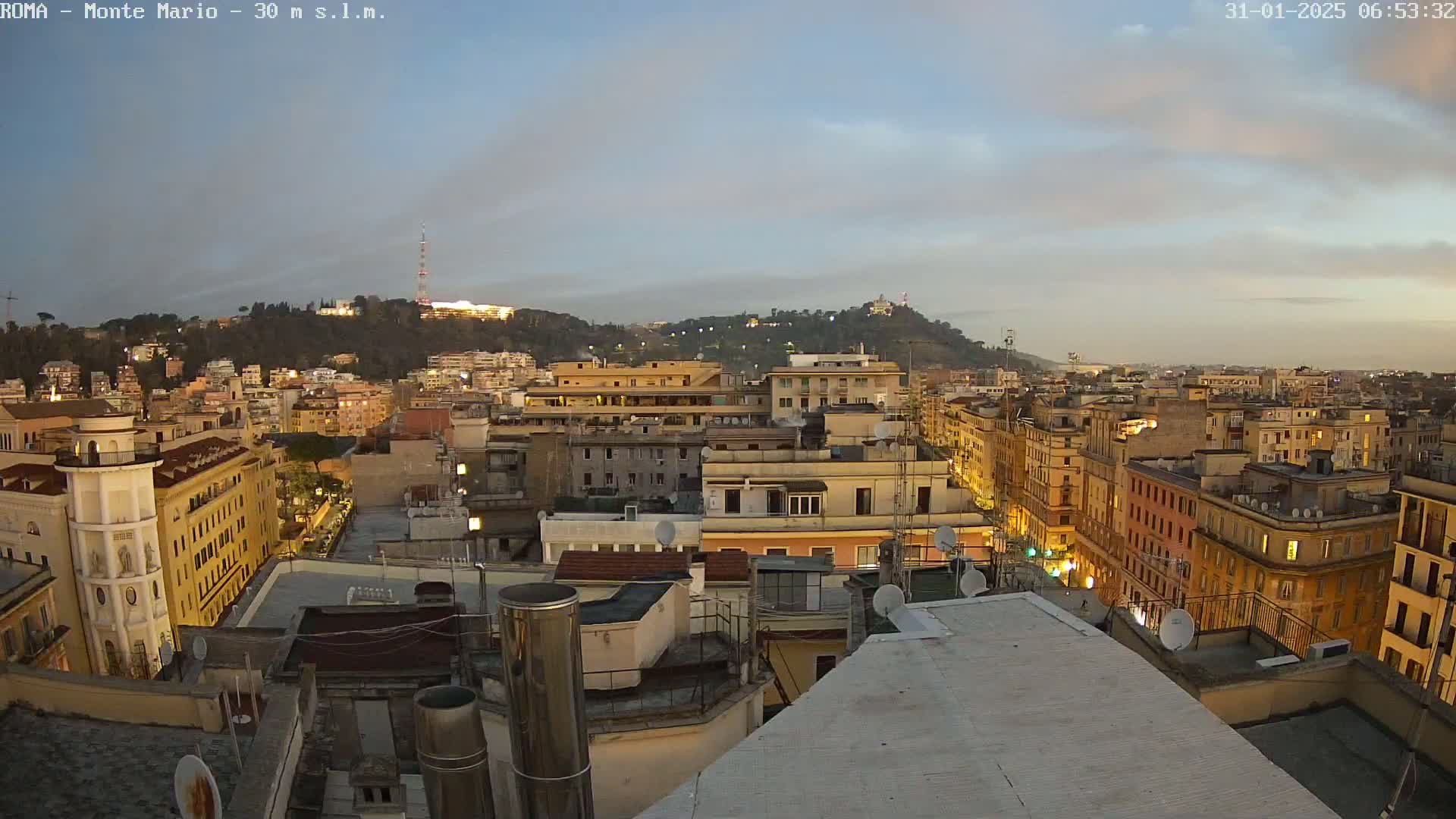 A twilight view of a cityscape, showing many buildings and a hill in the background under a partly cloudy sky.