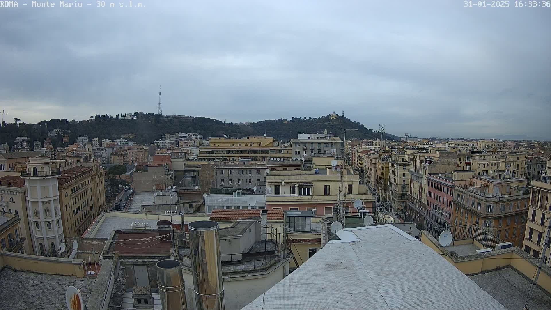 A high-angle view of a city under an overcast sky shows many buildings, a hill in the background, and a few antennas.