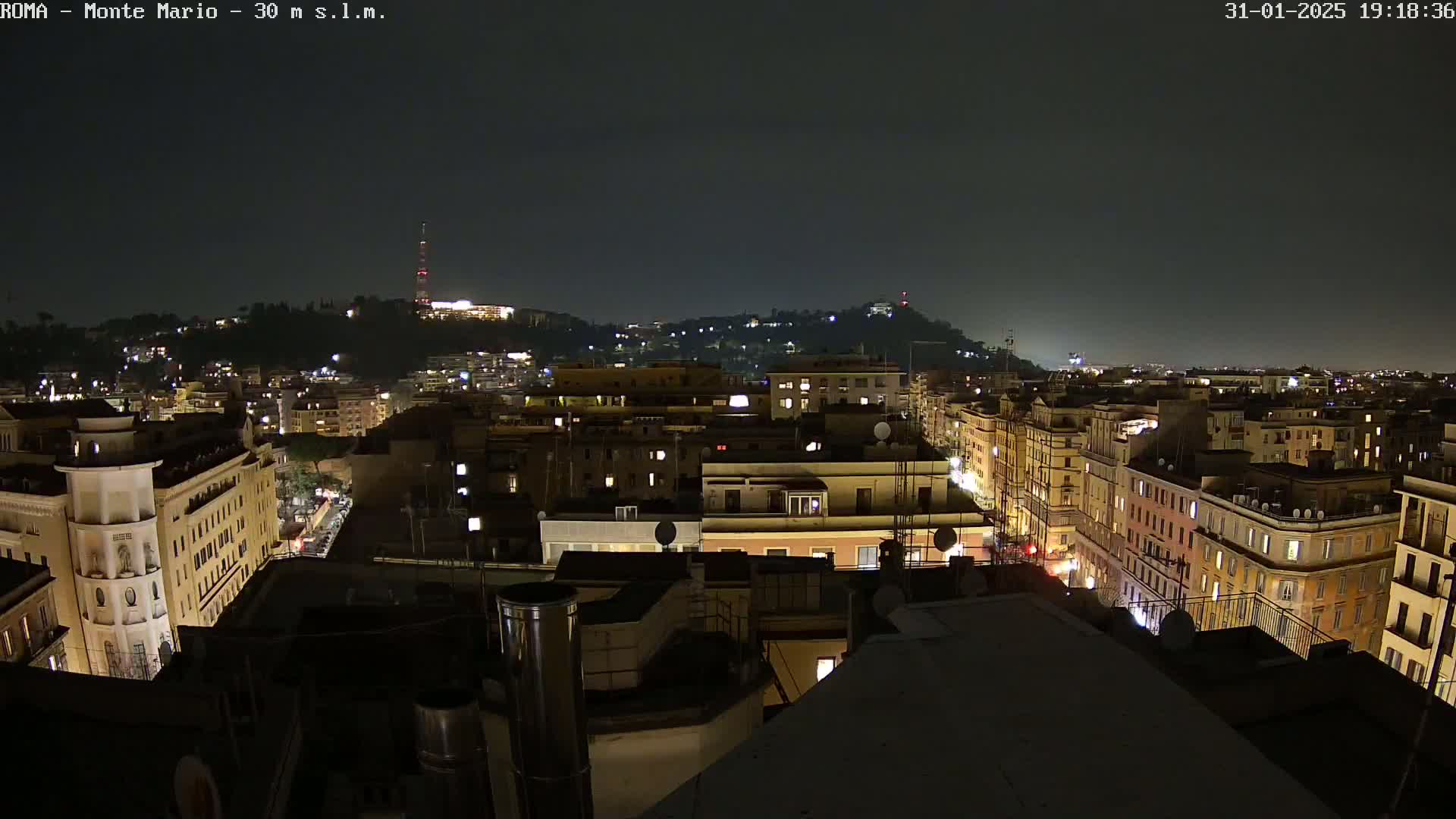 A nighttime, high-angle view of a city with illuminated buildings and streets, showing a hill in the background under a dark sky.