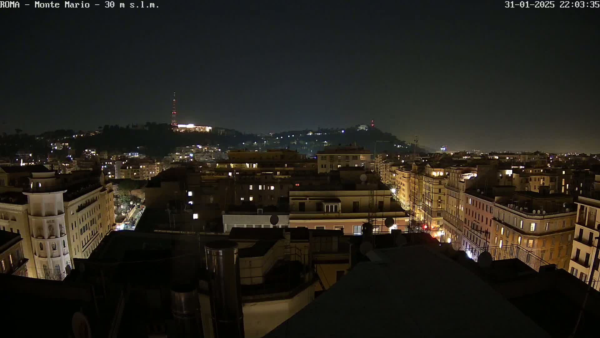 A nighttime view of a city illuminated by building lights, with hills in the distance under a clear, dark sky.