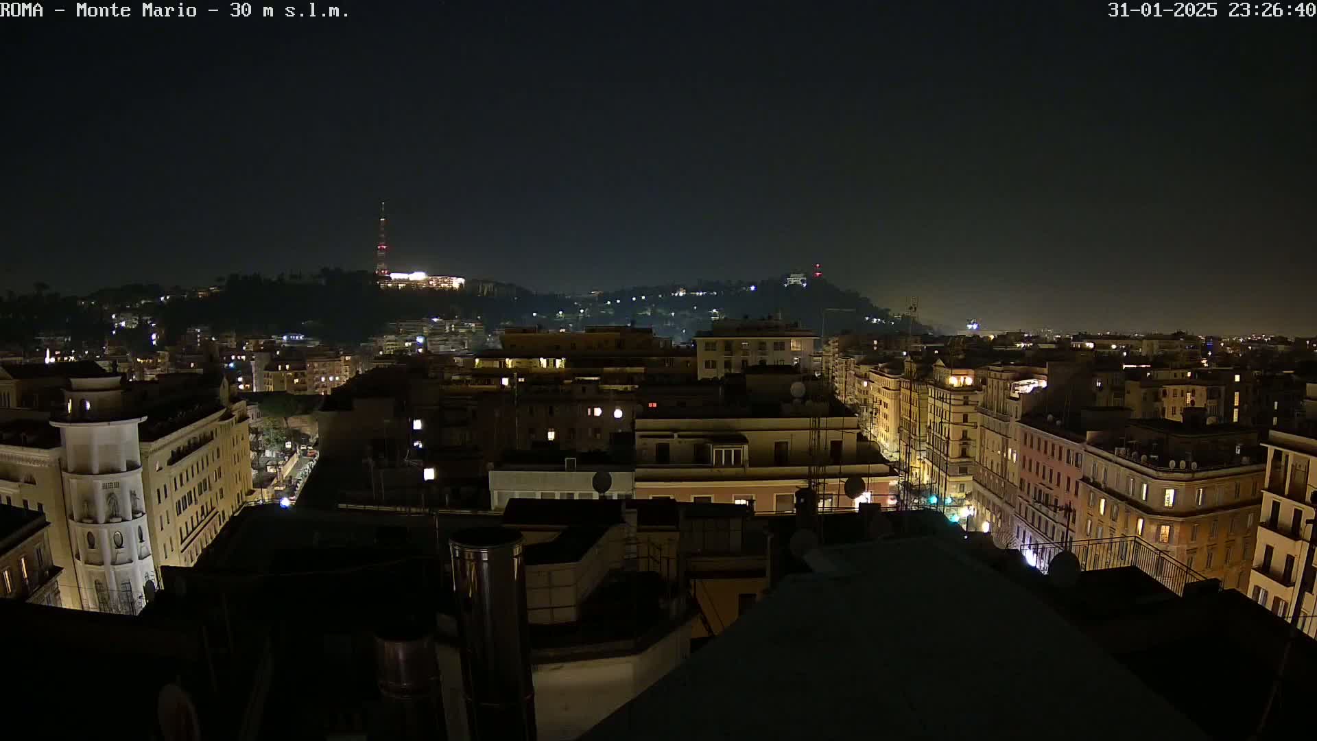A nighttime view of a city, with illuminated buildings and streets stretching towards a hill in the distance under a clear, dark sky.