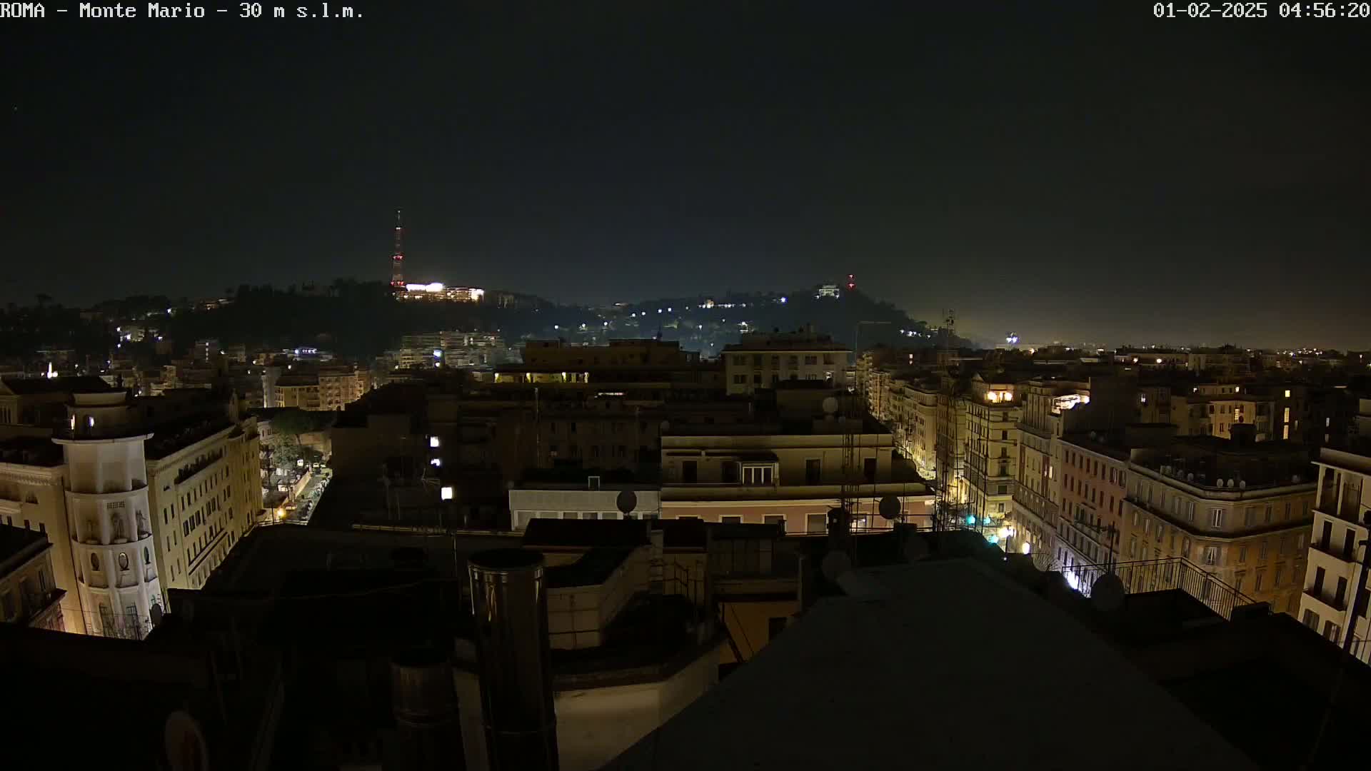 A nighttime, high-angle view of a city's rooftops and buildings, with a hill in the background dimly lit under a dark sky.