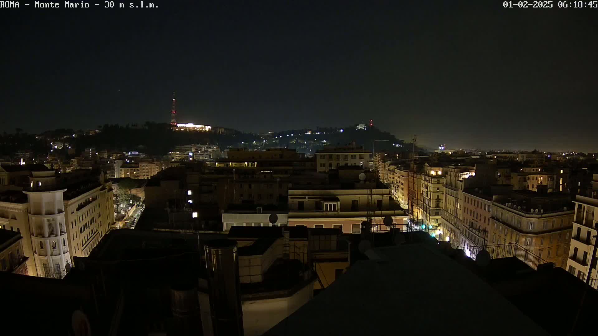 A nighttime view of a city, with dimly lit buildings and a hill in the distance under a clear, dark sky.