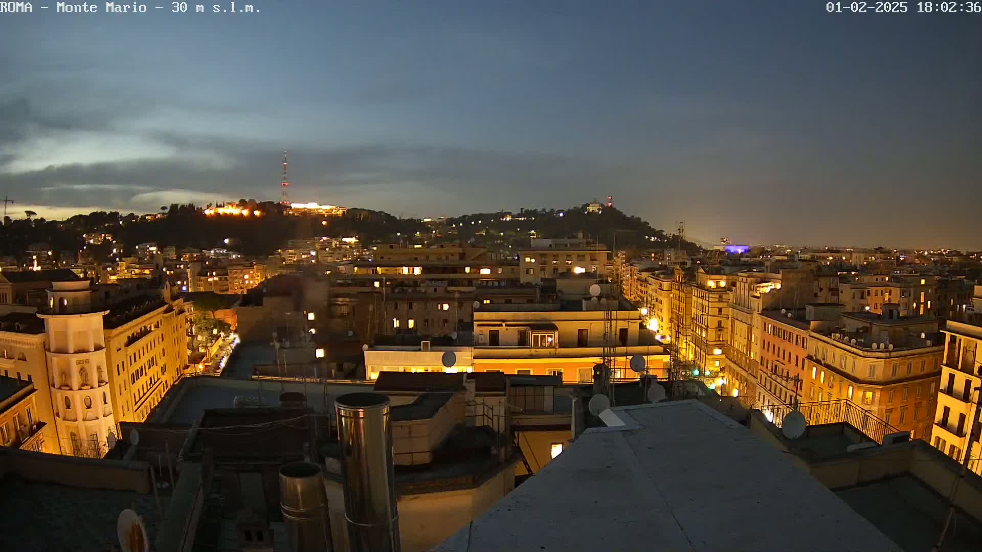 A nighttime view of a city, with illuminated buildings stretching to a hilltop in the distance under a mostly clear, dark sky.