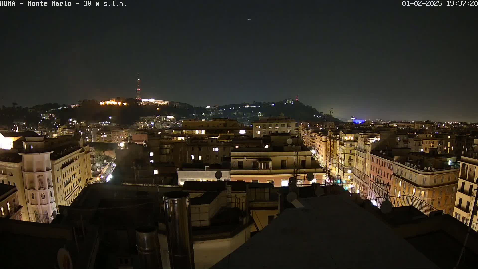 A nighttime, aerial view of a city shows many buildings with illuminated windows, extending to a hilltop in the distance under a clear, dark sky.
