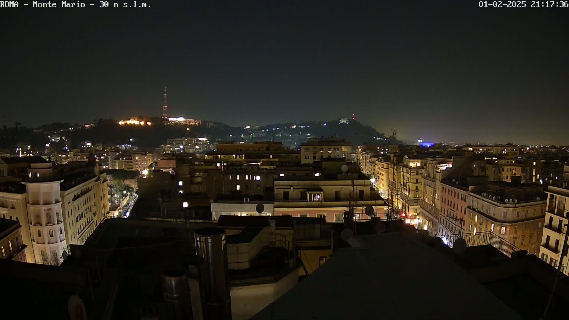A nighttime view of a city, showing numerous buildings illuminated with some streetlights visible, and a hill in the distance with more lights.