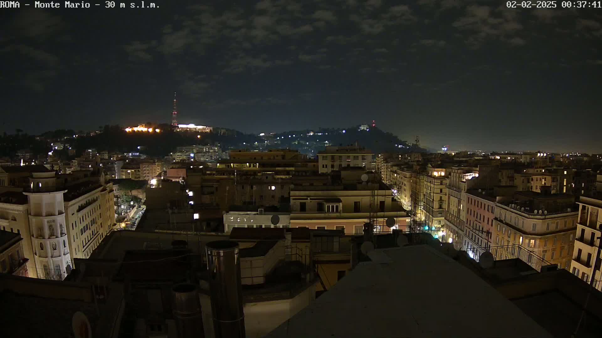 A nighttime view of a city with a hill in the background under a partly cloudy sky.