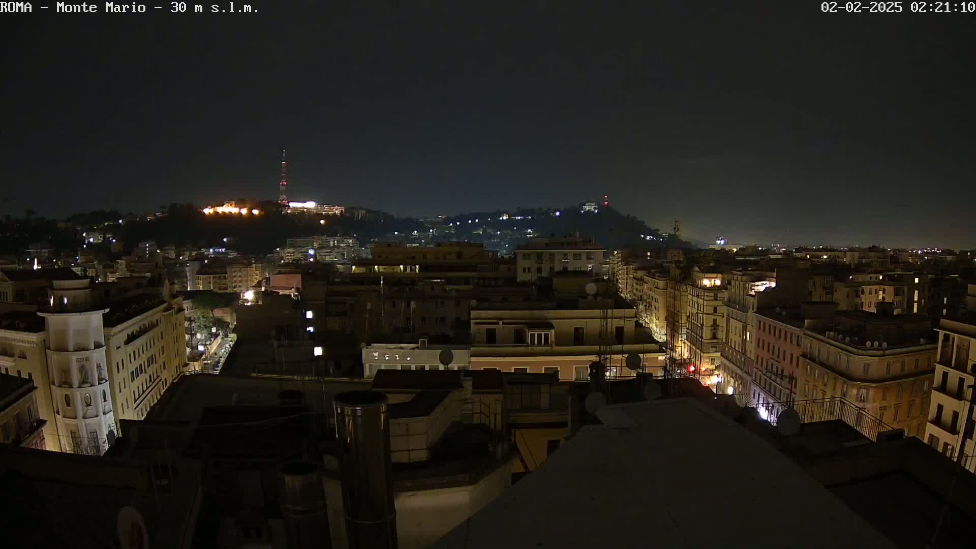 A nighttime cityscape view shows a hilly area with buildings illuminated, under a dark, clear sky.