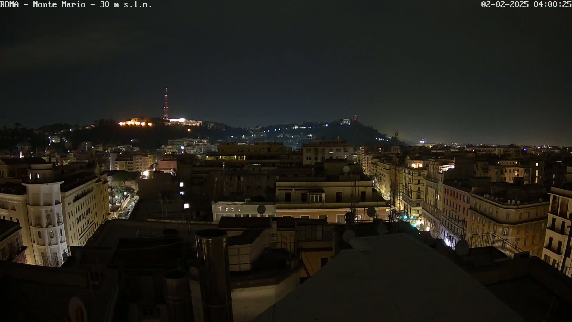 A nighttime view of a city, showing a hilltop in the distance with scattered lights, and city buildings with illuminated windows and streets below, under a dark sky.