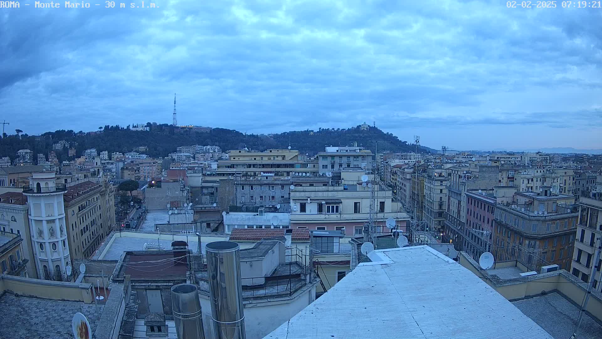 A cityscape under a cloudy, dusky sky shows numerous buildings, with a hill in the background.