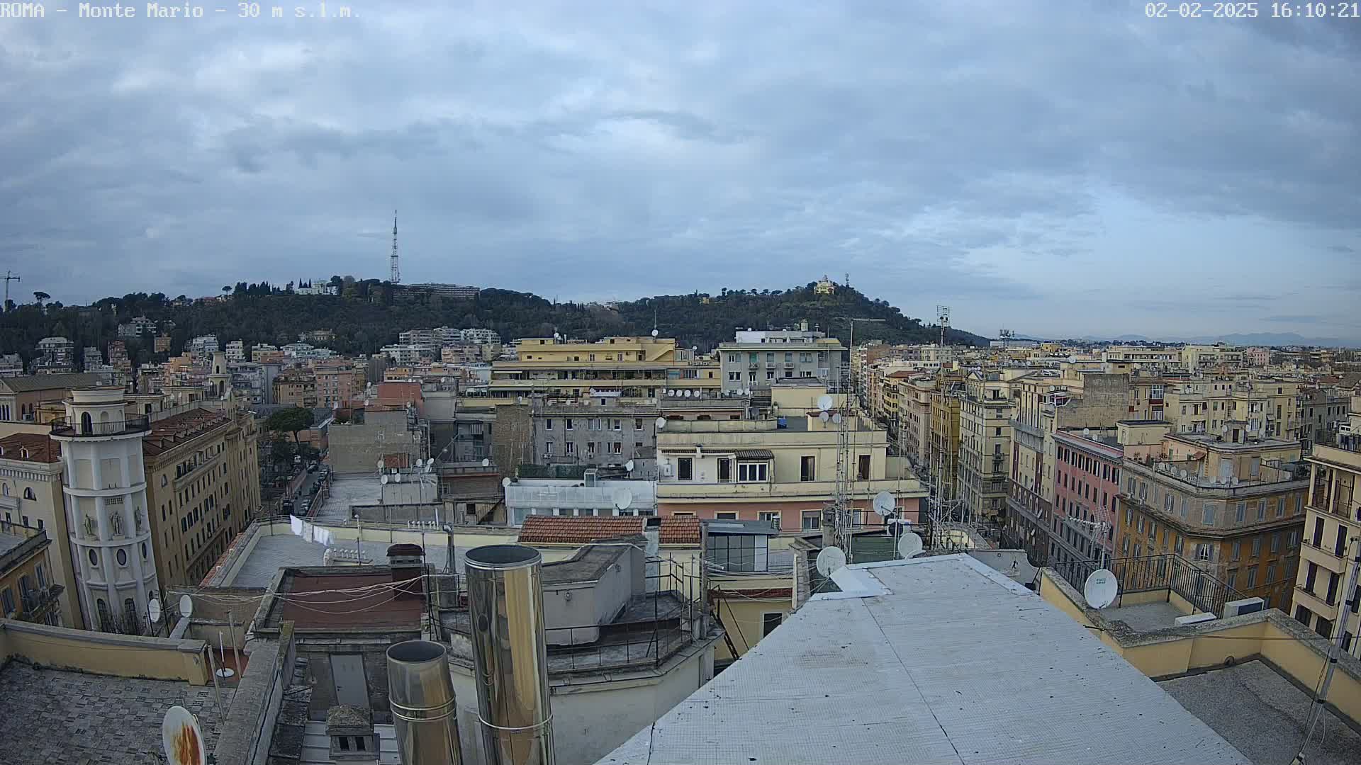 A panoramic view of a city under a cloudy sky shows many buildings, rooftops, and a hill in the distance.