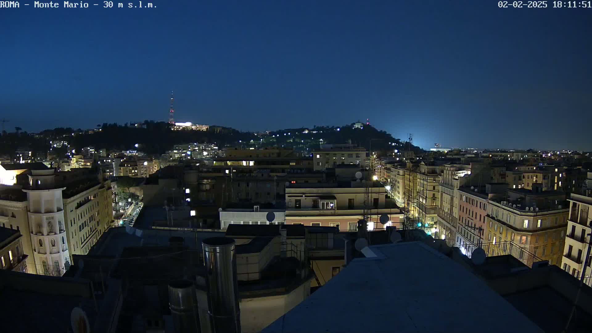 A nighttime cityscape of multi-story buildings, illuminated by streetlights and windows, extends to a hill in the distance under a clear, dark blue sky.