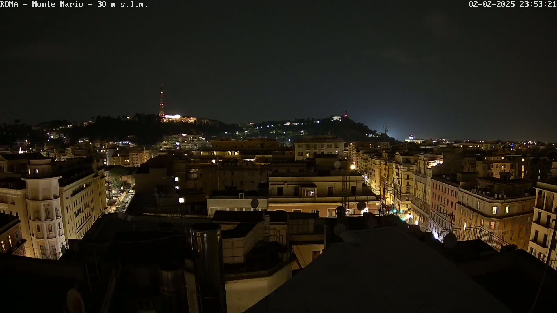 A nighttime, high-angle view of a city, showing many buildings with illuminated windows, extending to a hill in the distance under a dark sky.