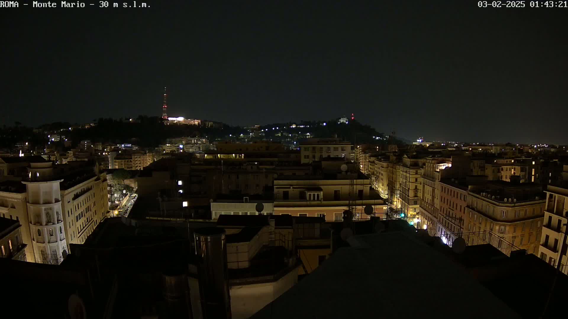 A nighttime view of a city, showing numerous buildings illuminated by streetlights and some interior lights, with a hill in the background also showing some lights.