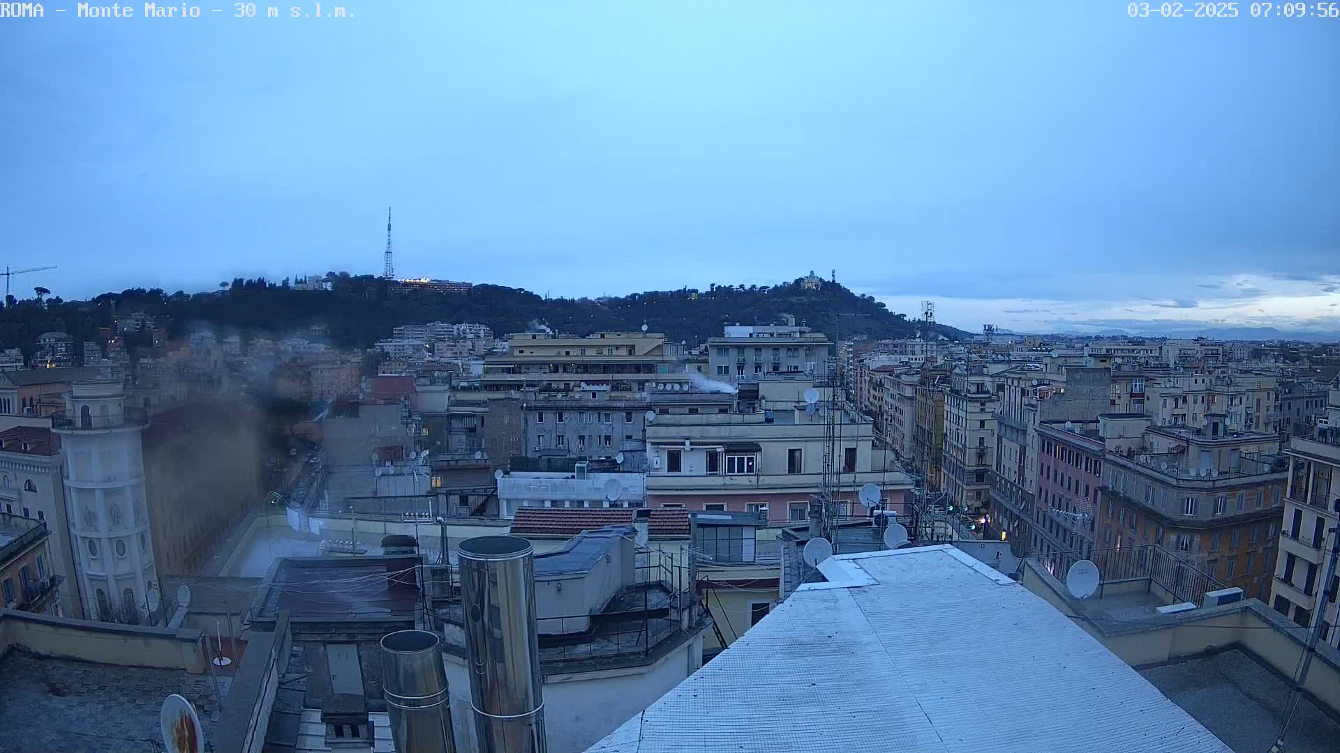 A view of a city skyline at dawn under a clear, light-blue sky, showing many buildings and rooftops, with a hill in the distance.