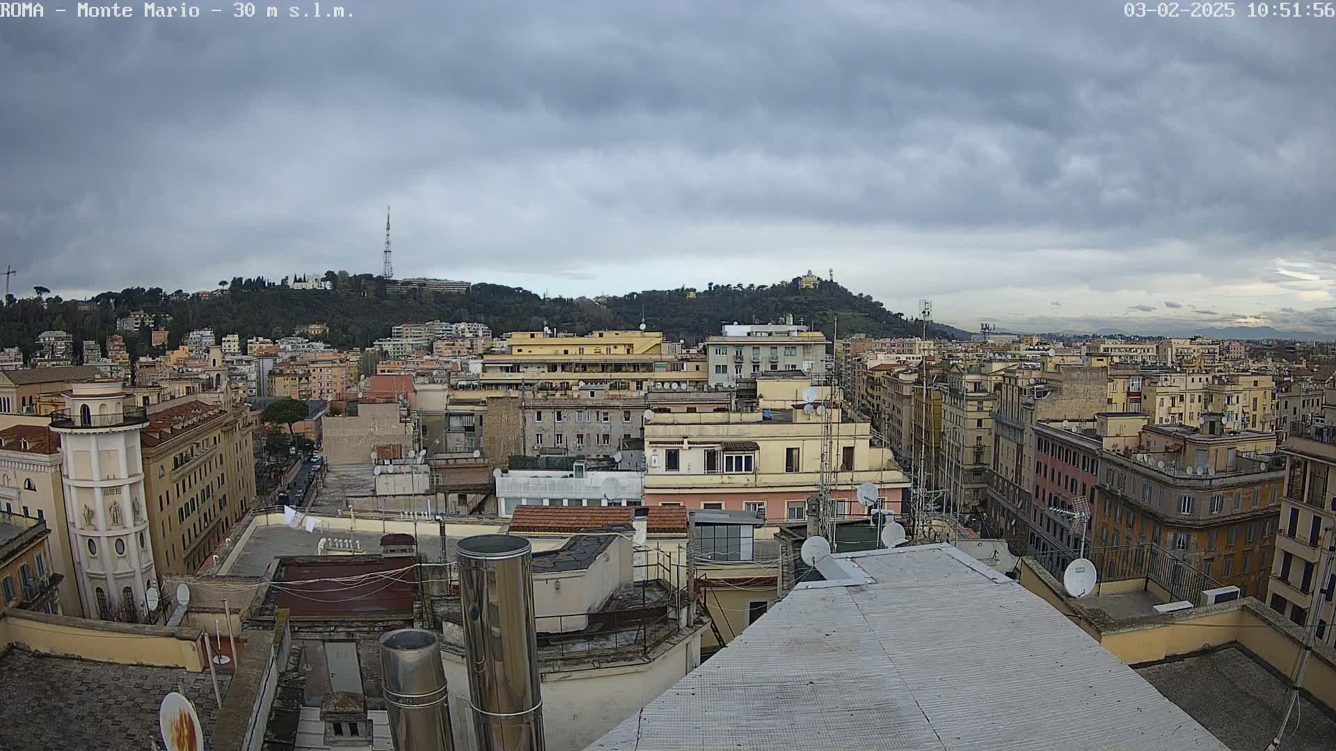 A city skyline under a cloudy sky shows many buildings, rooftops, and a hill in the distance.