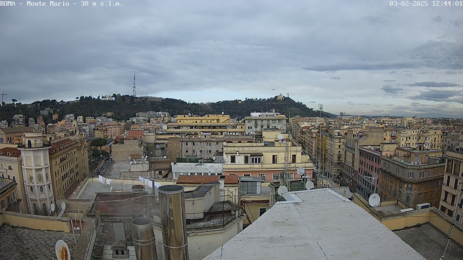 A panoramic view of a city under a cloudy sky shows numerous buildings, rooftops, and a hill in the distance.