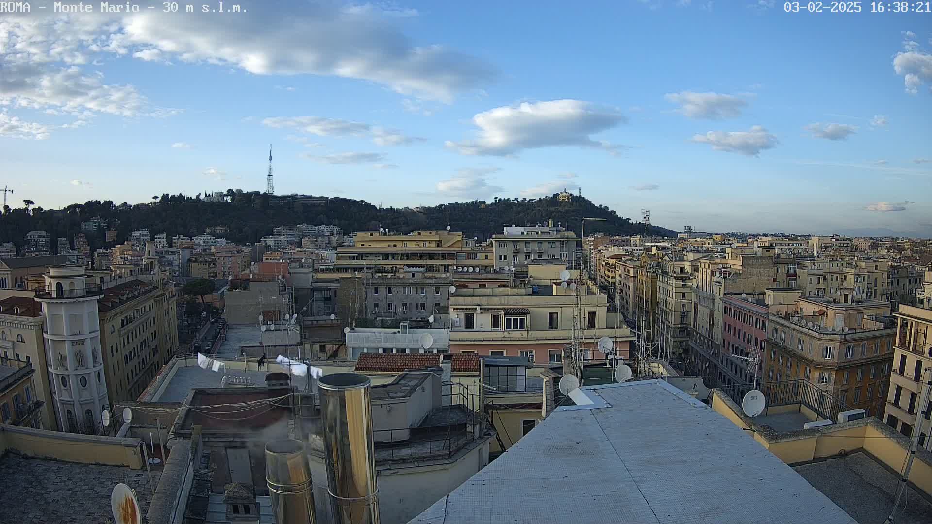 A mostly sunny day shows a sprawling cityscape of multi-story buildings, with a hill covered in trees in the background.