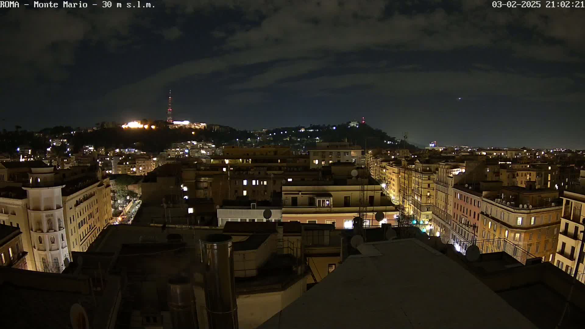 A nighttime cityscape view shows a hilltop in the distance, illuminated with lights, while the foreground displays a collection of city buildings with windows glowing from interior lights under a partly cloudy sky.