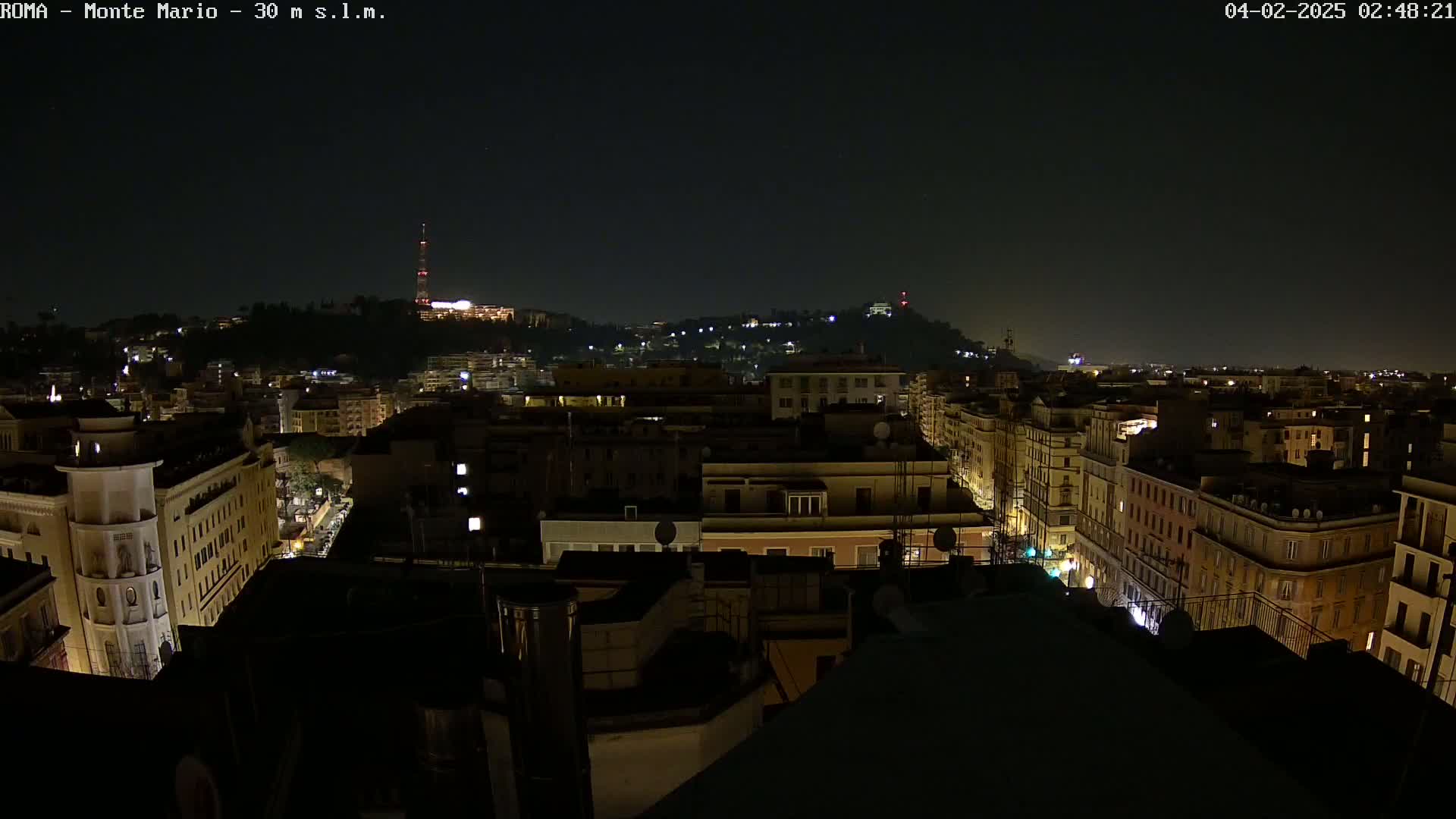 A nighttime view of a city, illuminated by streetlights and building lights, with a hill in the background under a clear, dark sky.