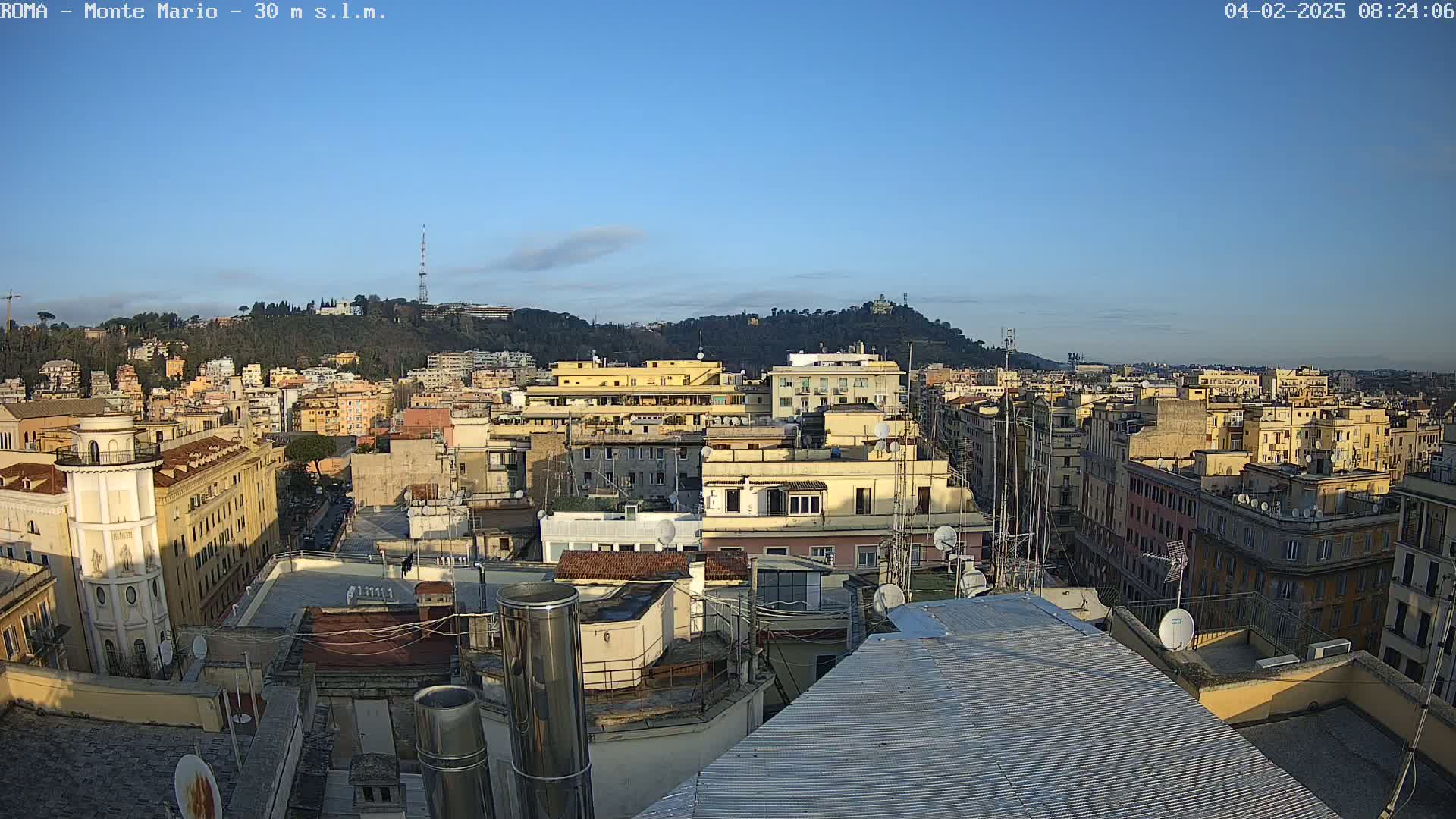 A rooftop view of a densely populated city under a clear blue sky shows numerous buildings, many with satellite dishes, extending to a distant hill.