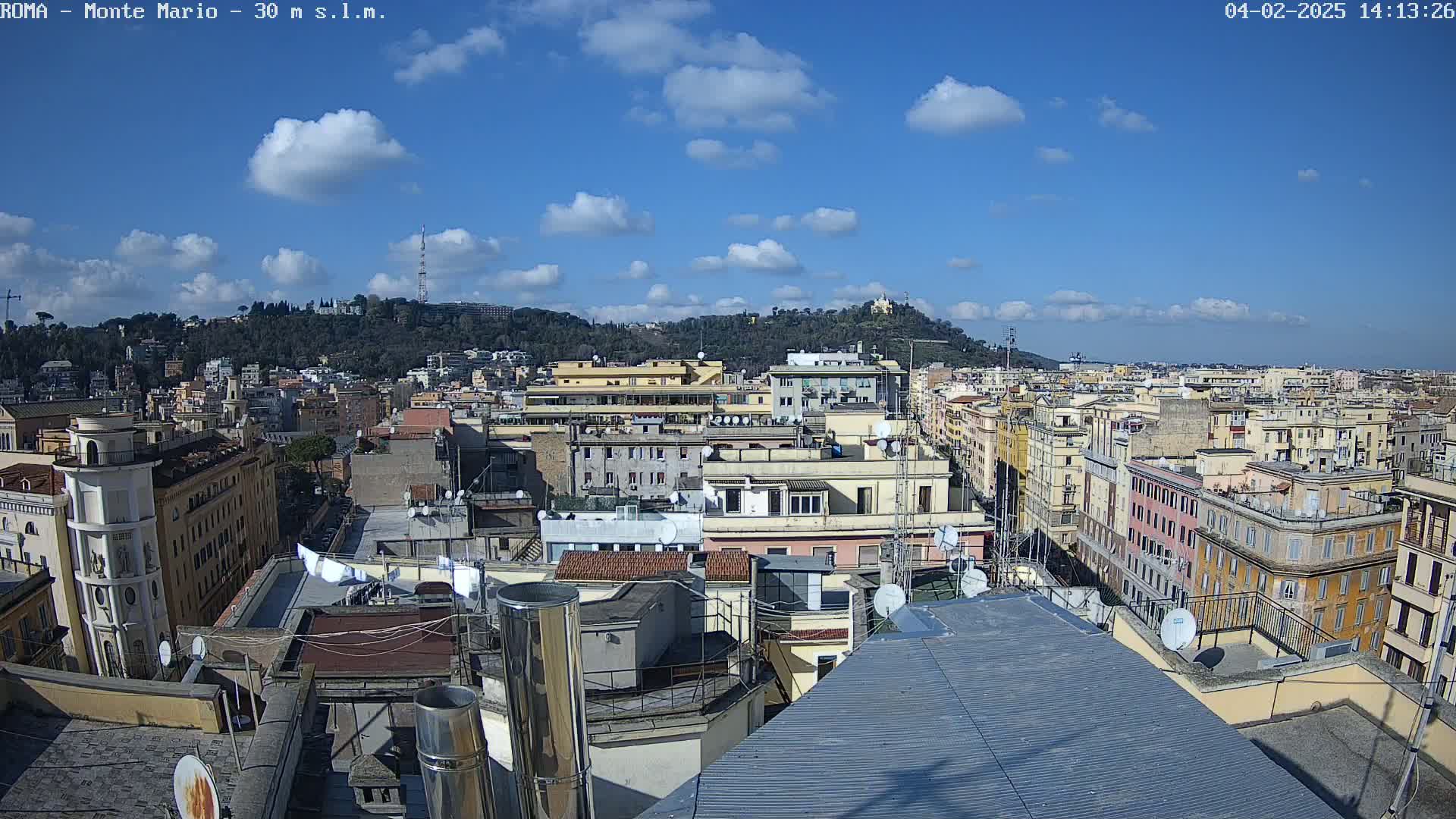 A mostly sunny cityscape view shows many buildings extending into the distance, with a hill in the background under a blue sky dotted with fluffy white clouds.