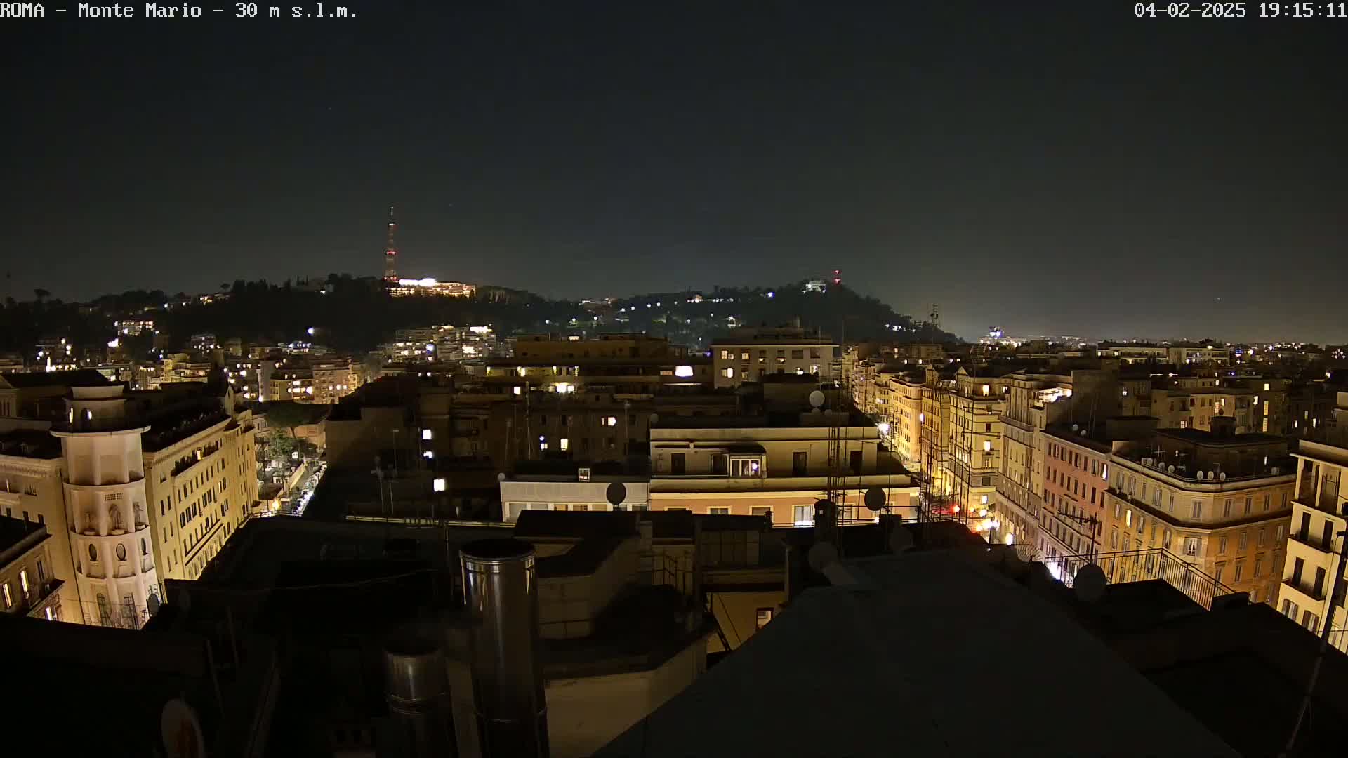 A nighttime view of a city, showing numerous buildings with illuminated windows, extending to a hill in the distance under a clear, dark sky.