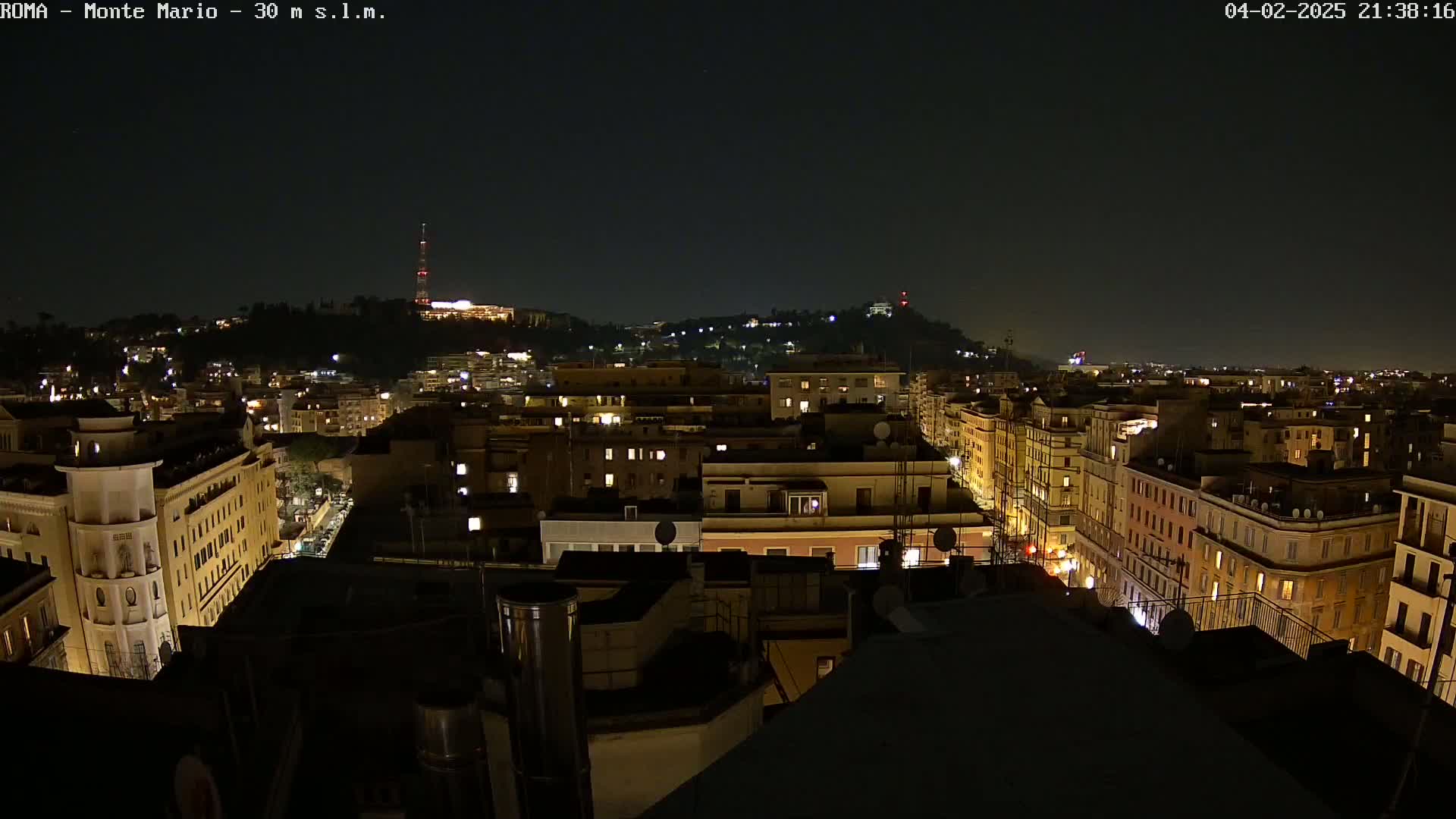 A nighttime cityscape of buildings with illuminated windows stretches out under a clear, dark sky, with a hill in the distance also showing lights.