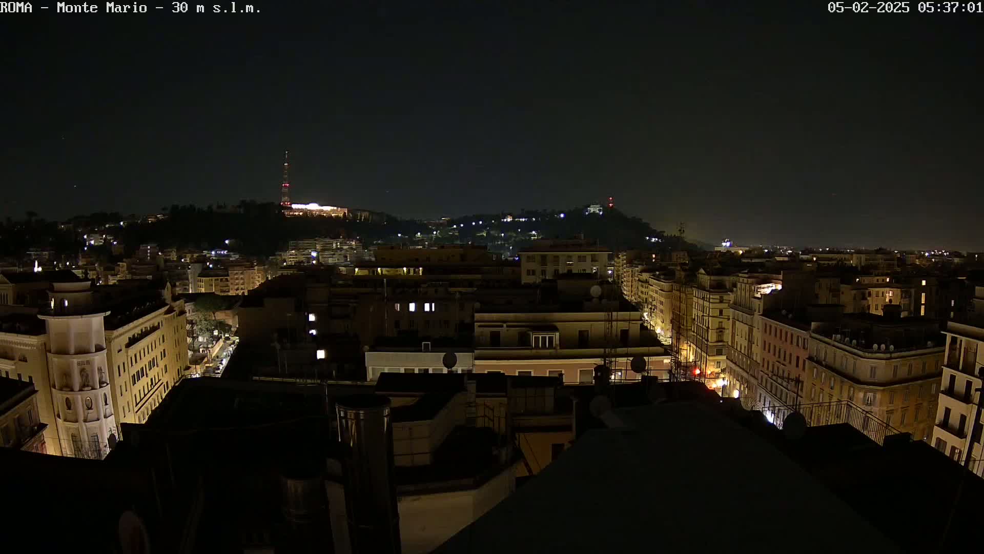 A nighttime view of a city, showing brightly lit buildings and streets, with a hill in the distance under a dark, clear sky.