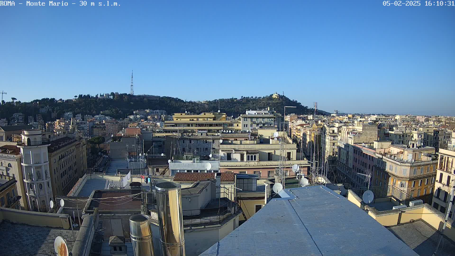 A high-angle, long shot on a clear, sunny day shows a cityscape of many densely packed buildings, with a hill in the background.
