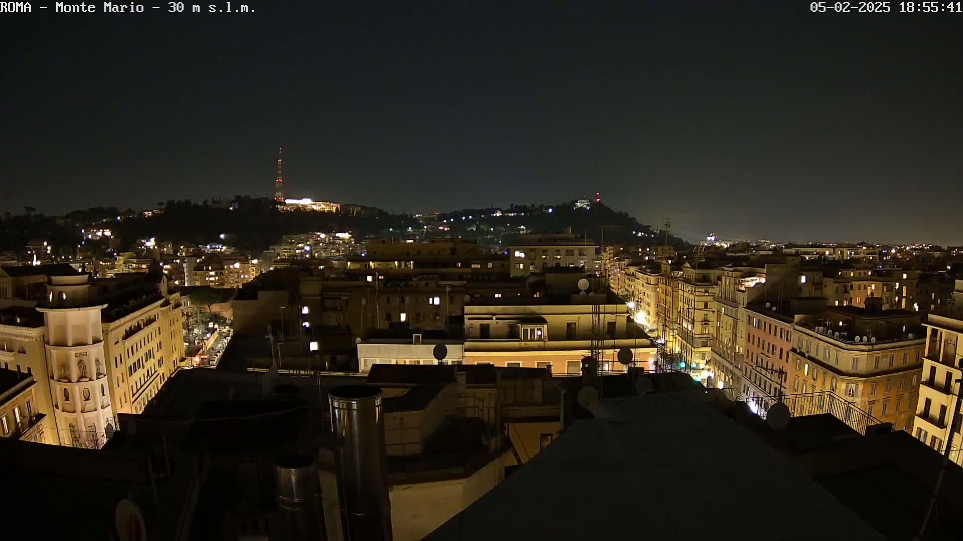 A nighttime view of a city, illuminated by lights from buildings and streets, with a hill in the background under a clear, dark sky.