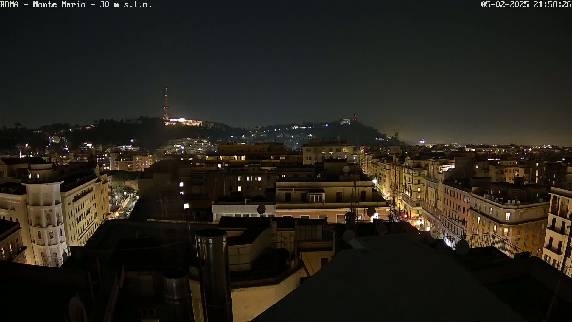 A nighttime view of a city, showing numerous buildings illuminated with warm-toned lights, and a hill in the distance under a clear, dark sky.