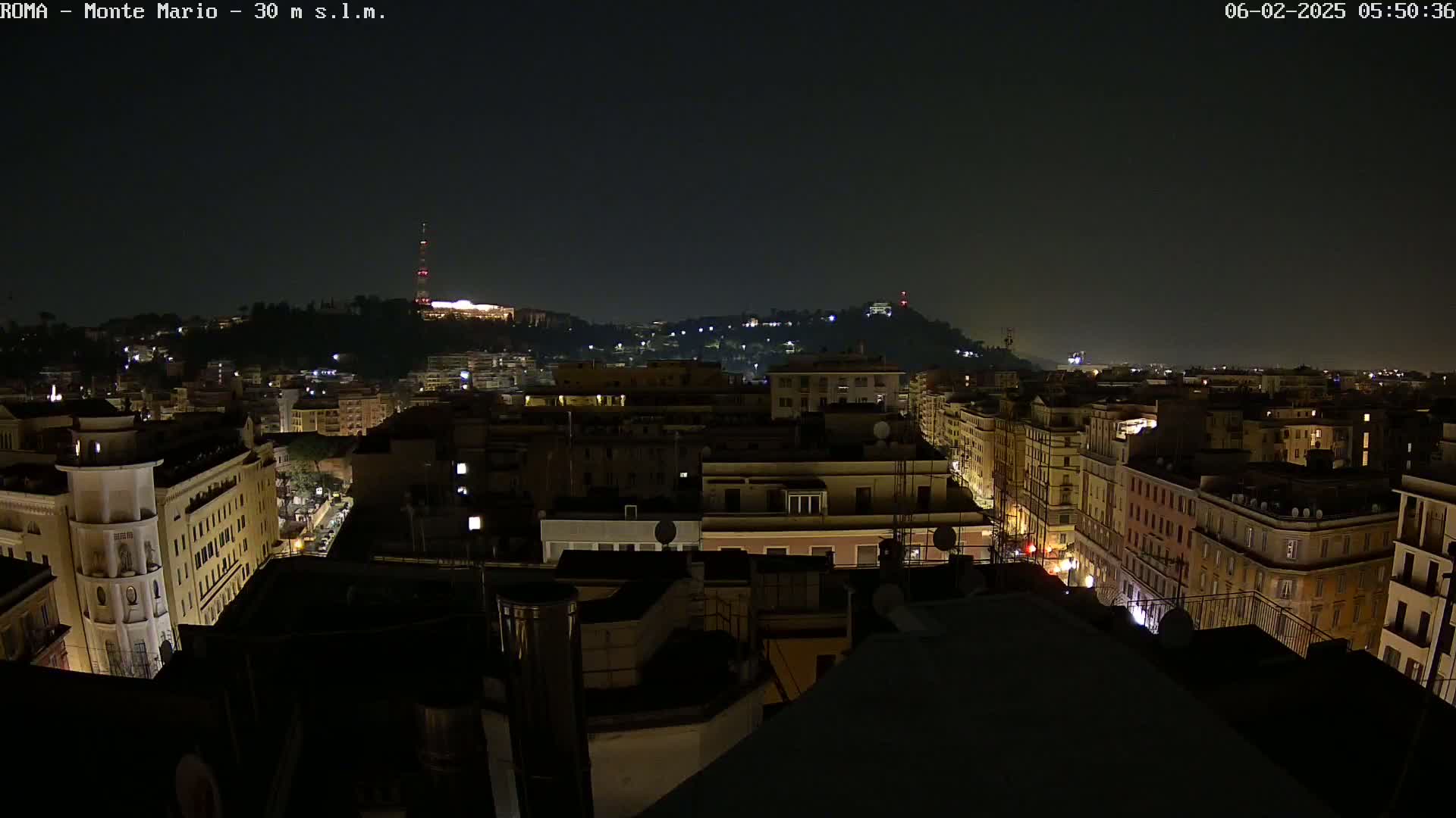 A nighttime view of a city skyline, with dimly lit buildings and streets extending to a hill in the distance under a dark sky.