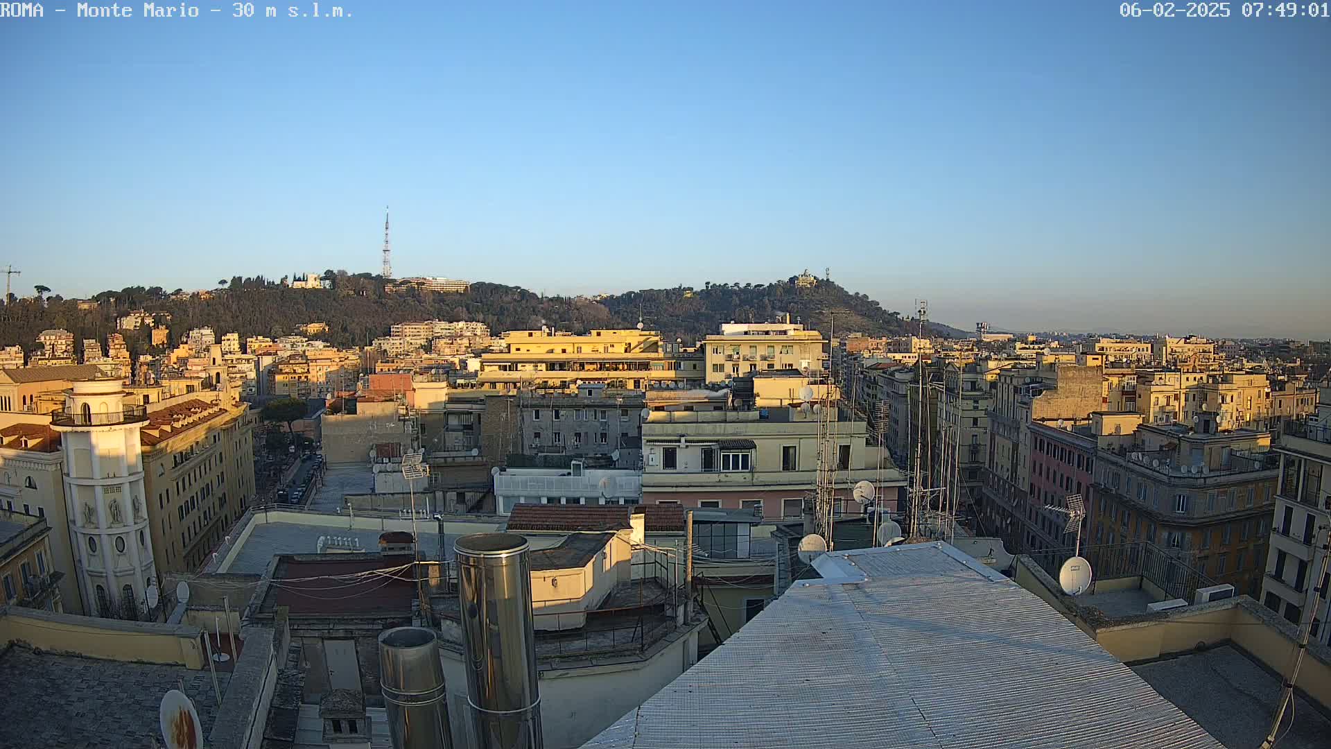 A rooftop view of a city in the early morning light, under a clear blue sky, shows numerous buildings extending to a hill in the distance.