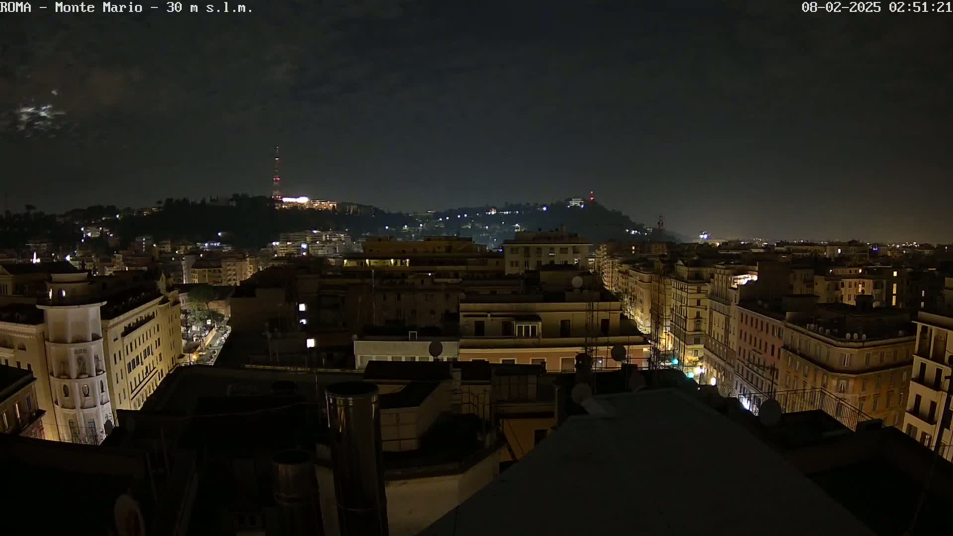 A nighttime view of a city, showing a hilltop in the distance under a mostly clear sky, with dimly lit buildings and streets visible below.