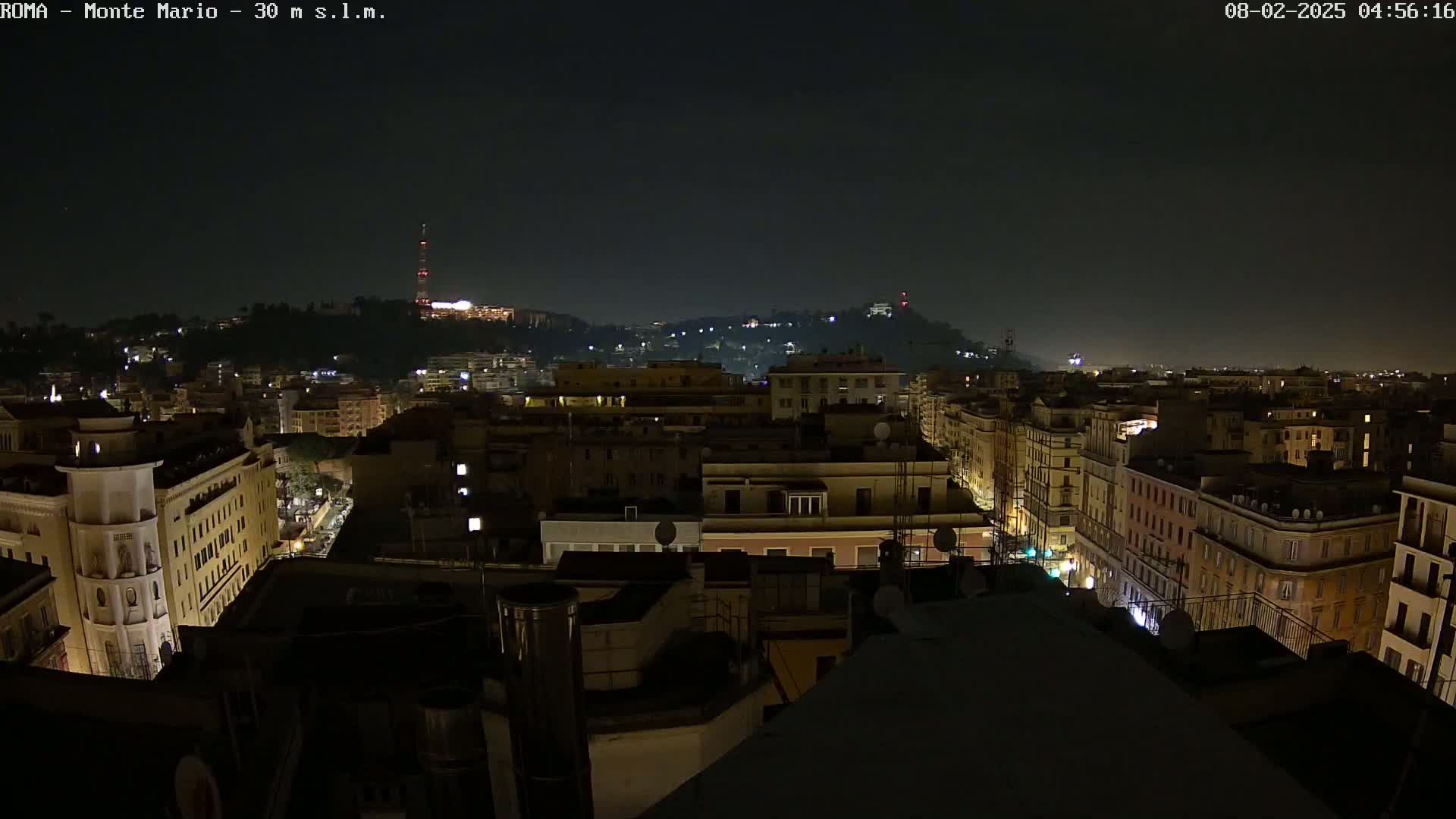A nighttime view of a city skyline, showing numerous buildings illuminated by streetlights and other lights, with a hill in the background under a dark, clear sky.
