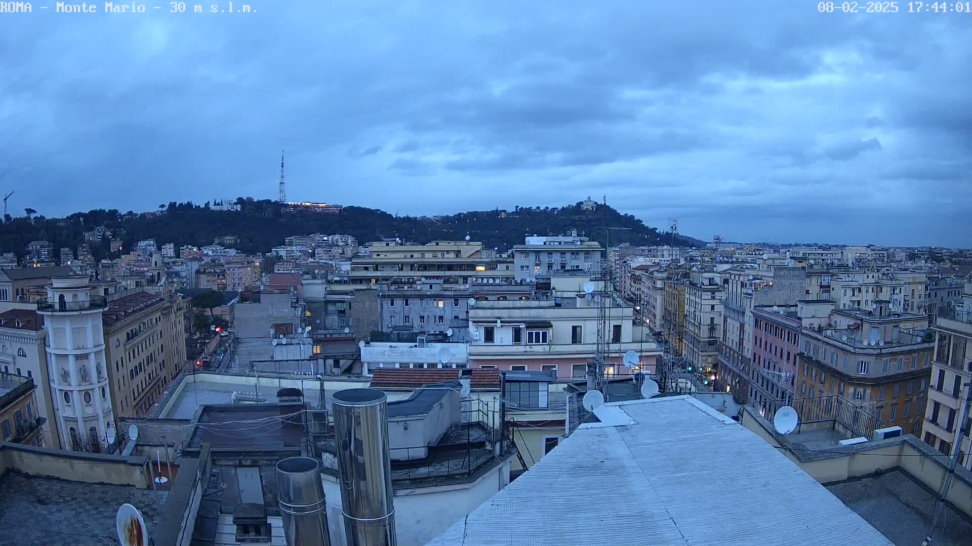 A view from a rooftop shows a cityscape at twilight under a cloudy sky, with a hill in the background.