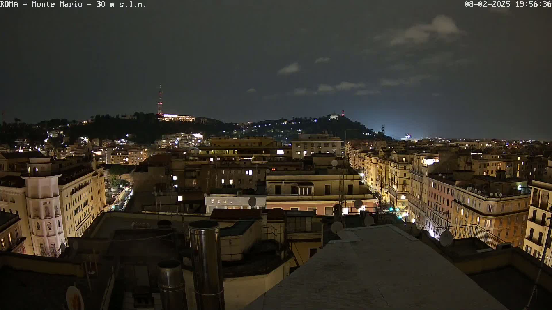 A nighttime view of a city with a hill in the background under a partly cloudy sky.