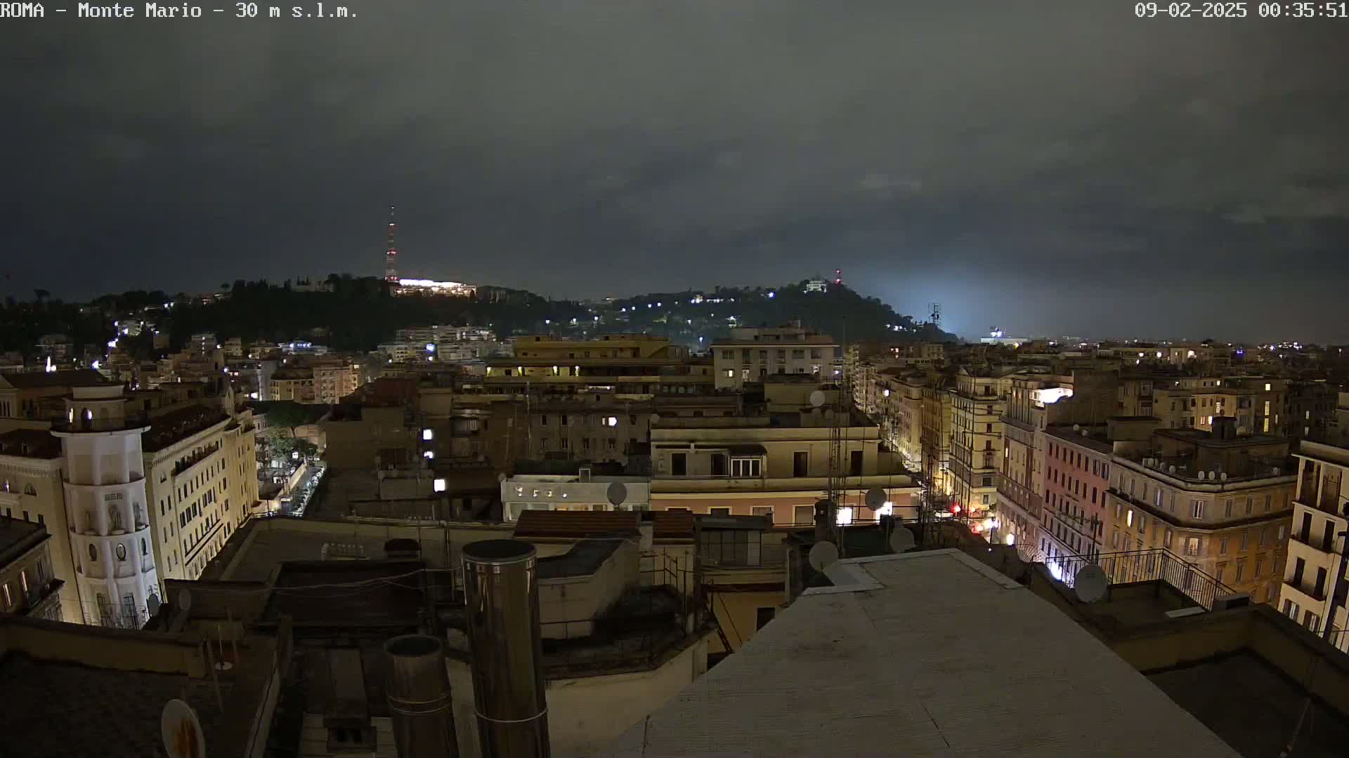 A nighttime view of a city with a hill in the background under a dark, cloudy sky.