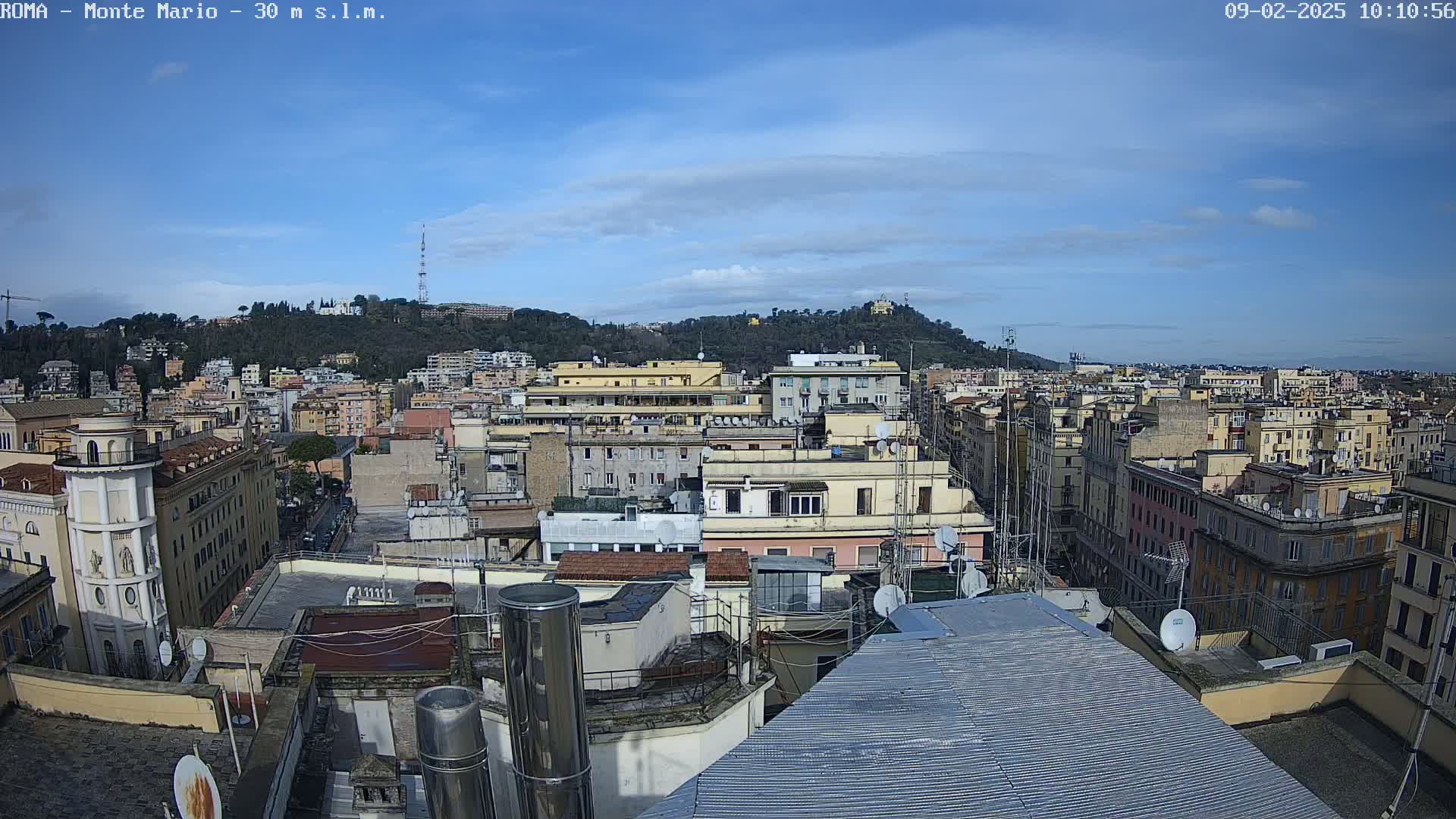 A mostly sunny day reveals a cityscape of densely packed buildings, with a hill in the background featuring trees and structures.