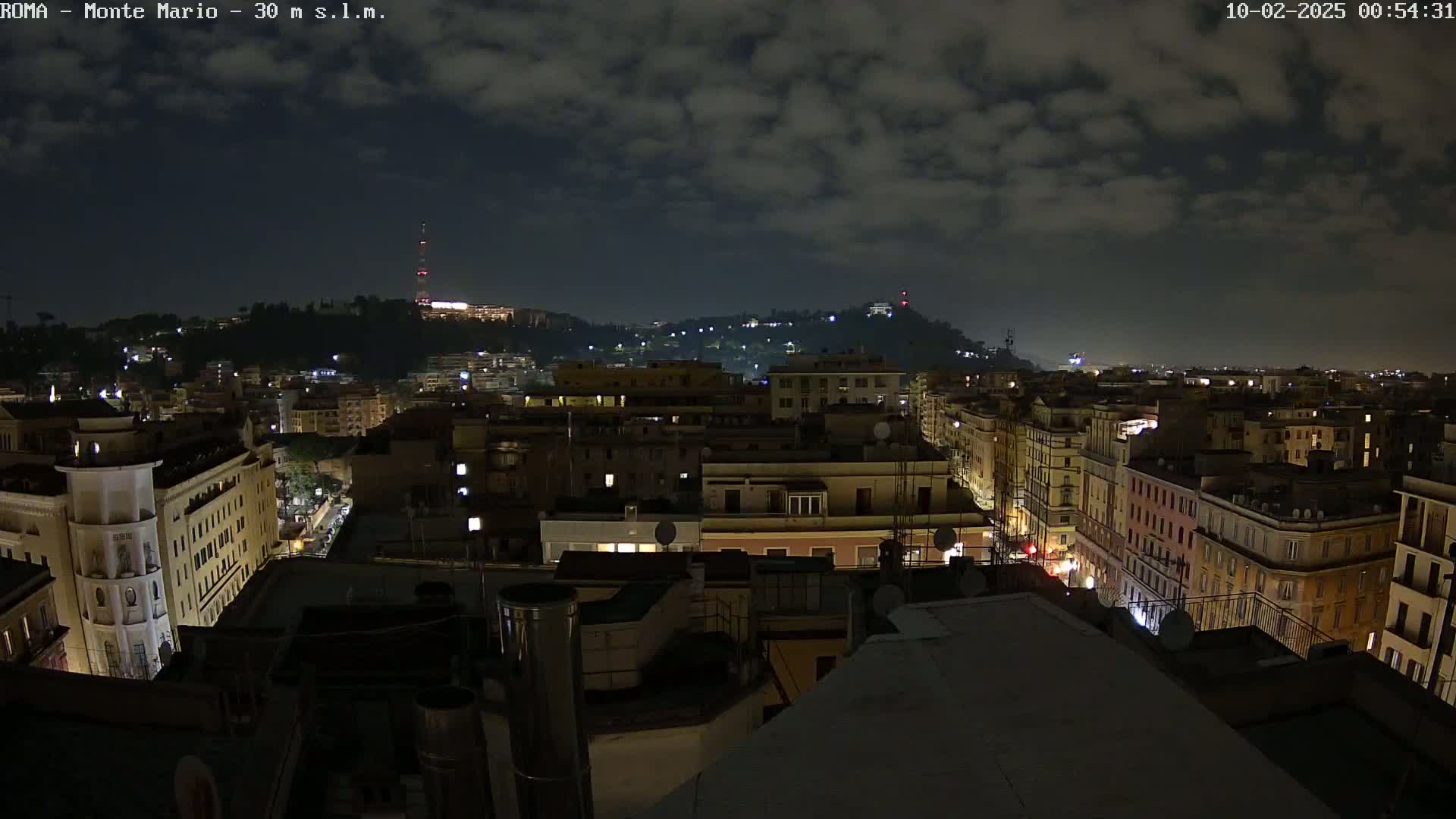 A nighttime view of a city, under a cloudy sky, shows numerous buildings illuminated, with a hill in the background also showing some lights.