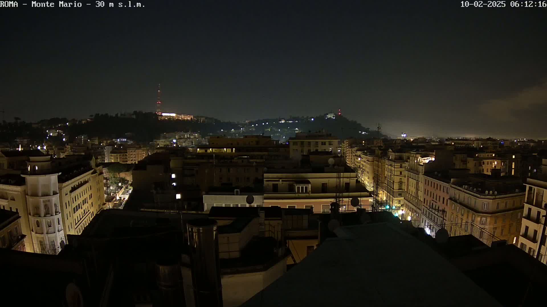 A nighttime view of a city shows buildings illuminated by streetlights and some interior lights, with a hill in the background under a dark, slightly hazy sky.