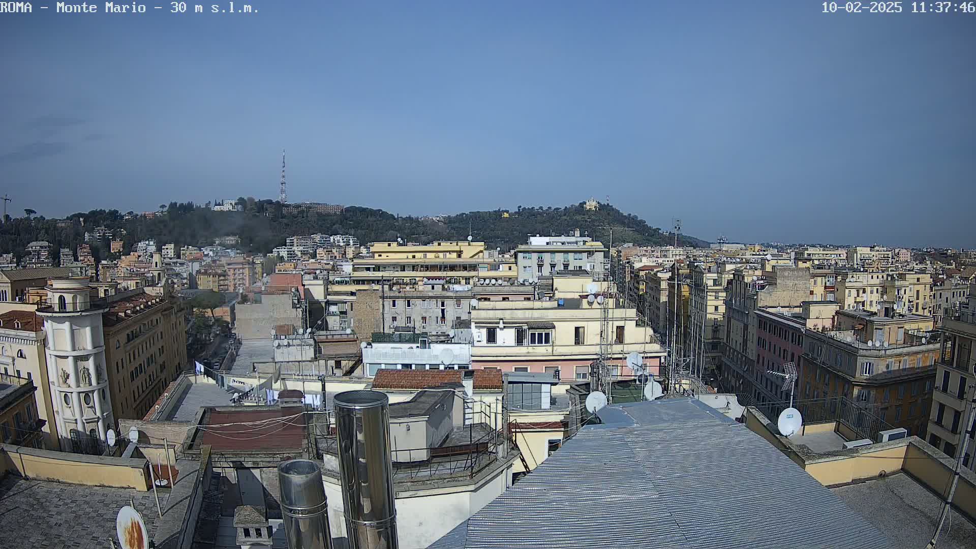 A clear, sunny day reveals a sprawling cityscape of densely packed buildings, with a hill in the background dotted with greenery and structures.