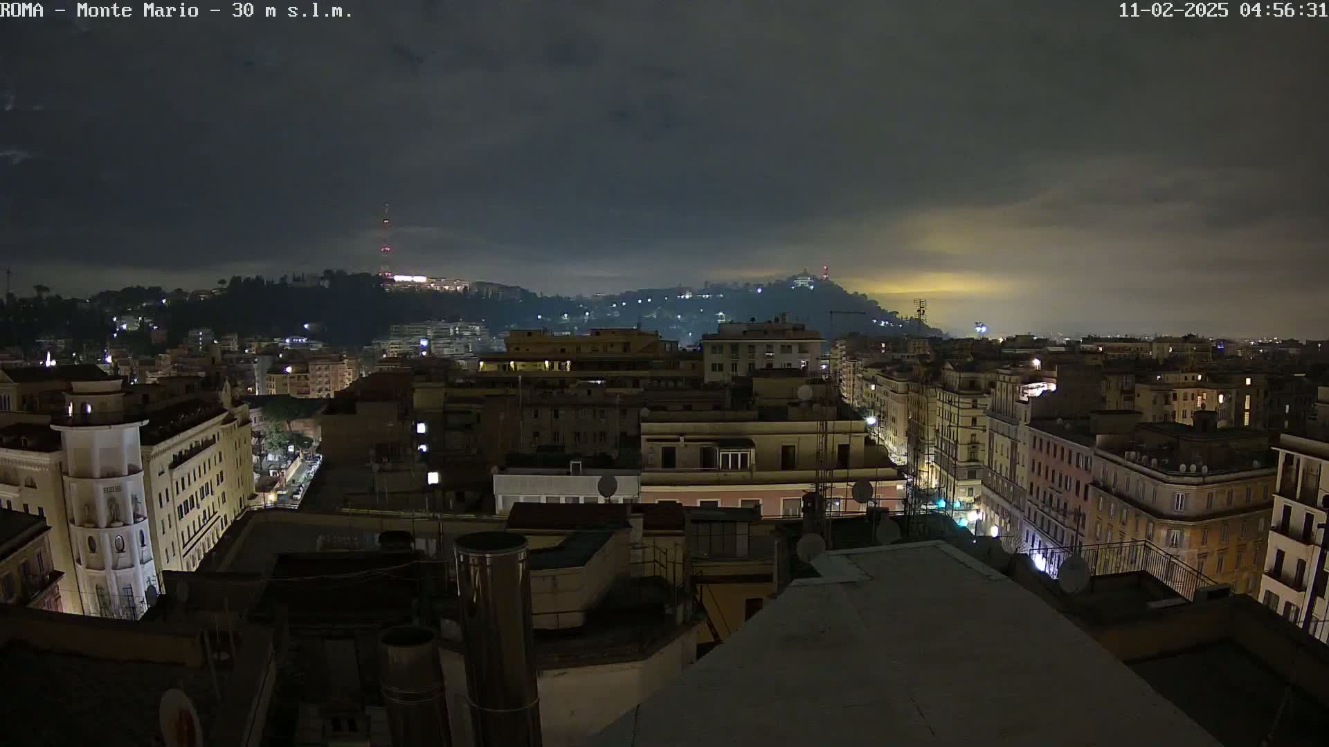 A nighttime view of a city, under a cloudy sky, shows a densely packed area of buildings lit with streetlights and windows.