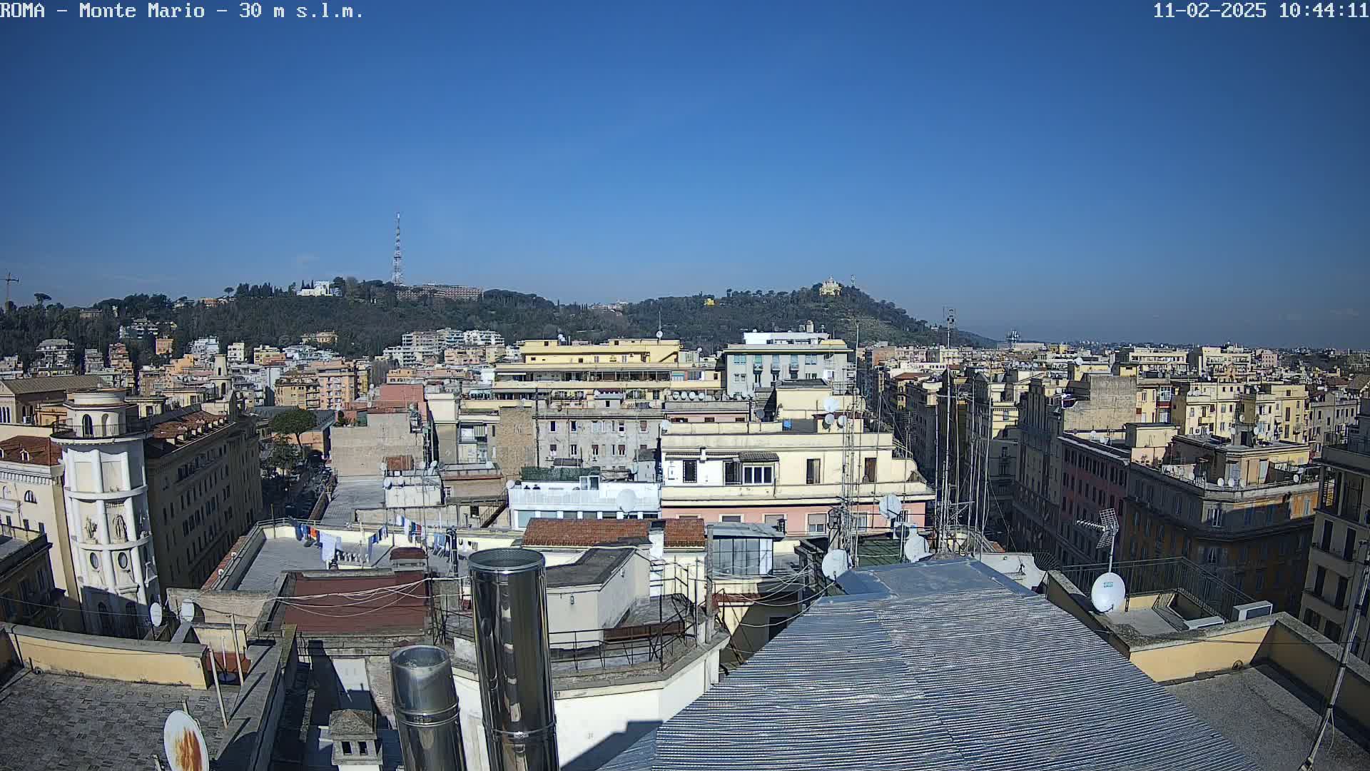 A rooftop view of a dense city under a clear blue sky shows numerous buildings extending to a distant hill.