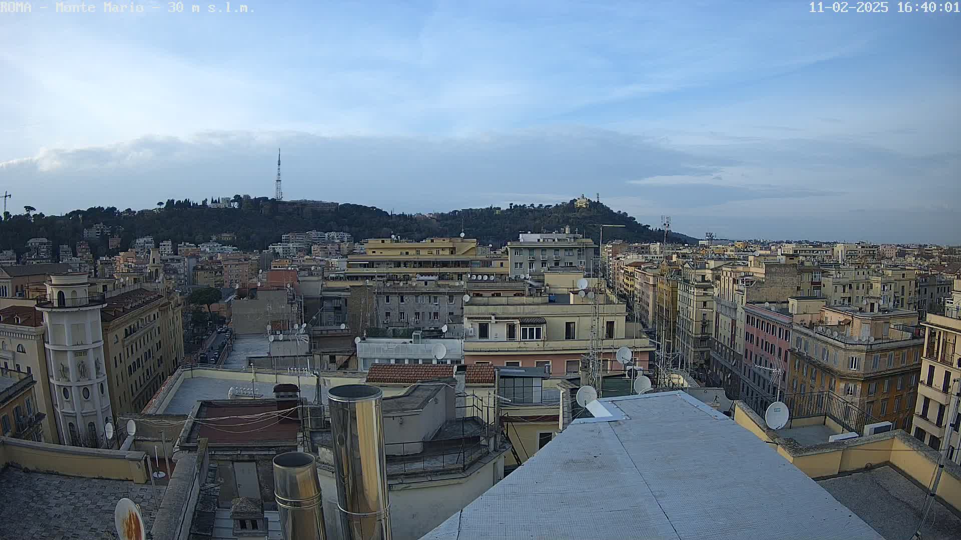 An aerial view of a city under a mostly clear sky shows many buildings extending to a hill in the distance.
