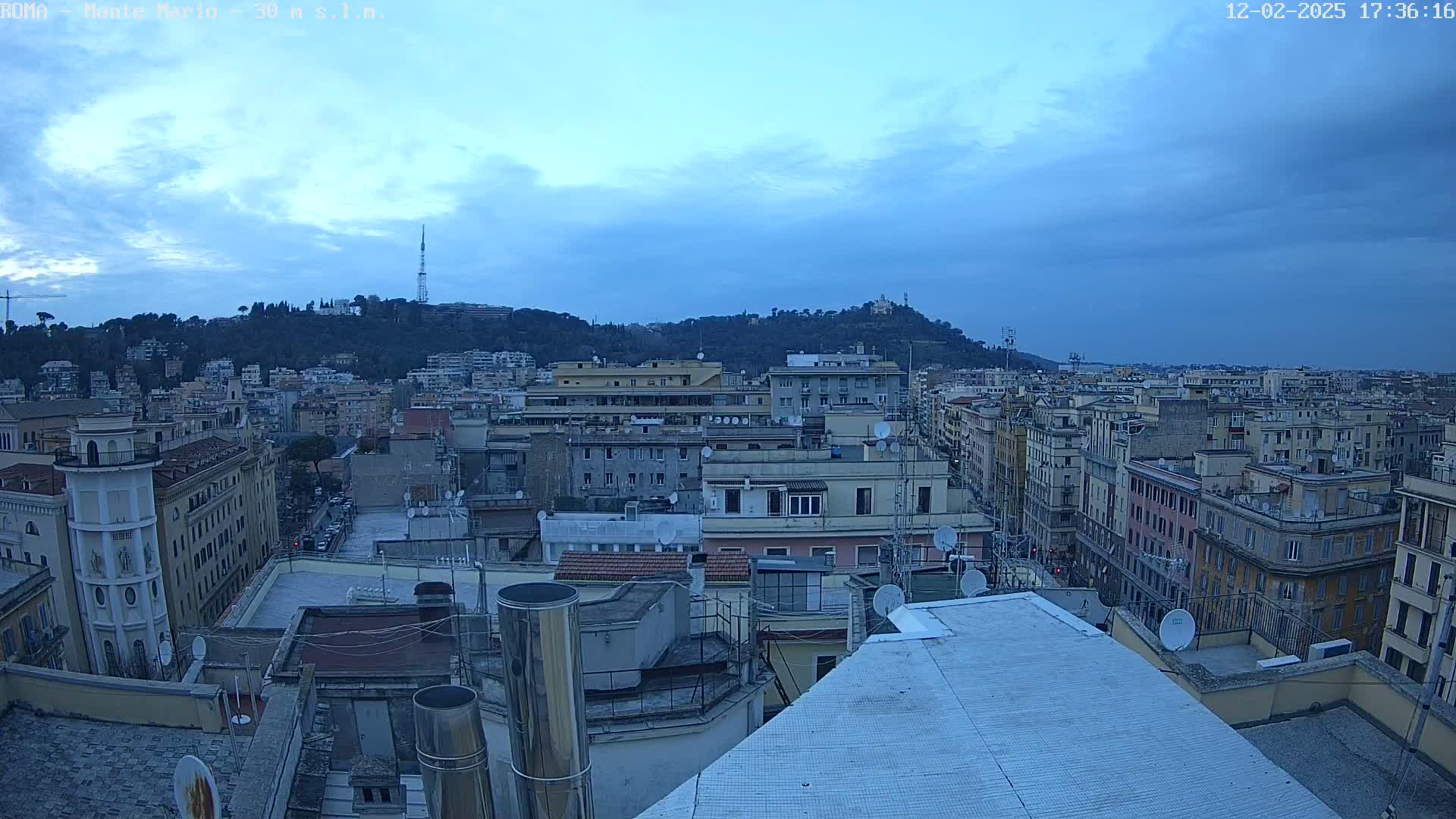 A mostly cloudy, twilight cityscape view shows numerous buildings and rooftops, with a hill in the background.