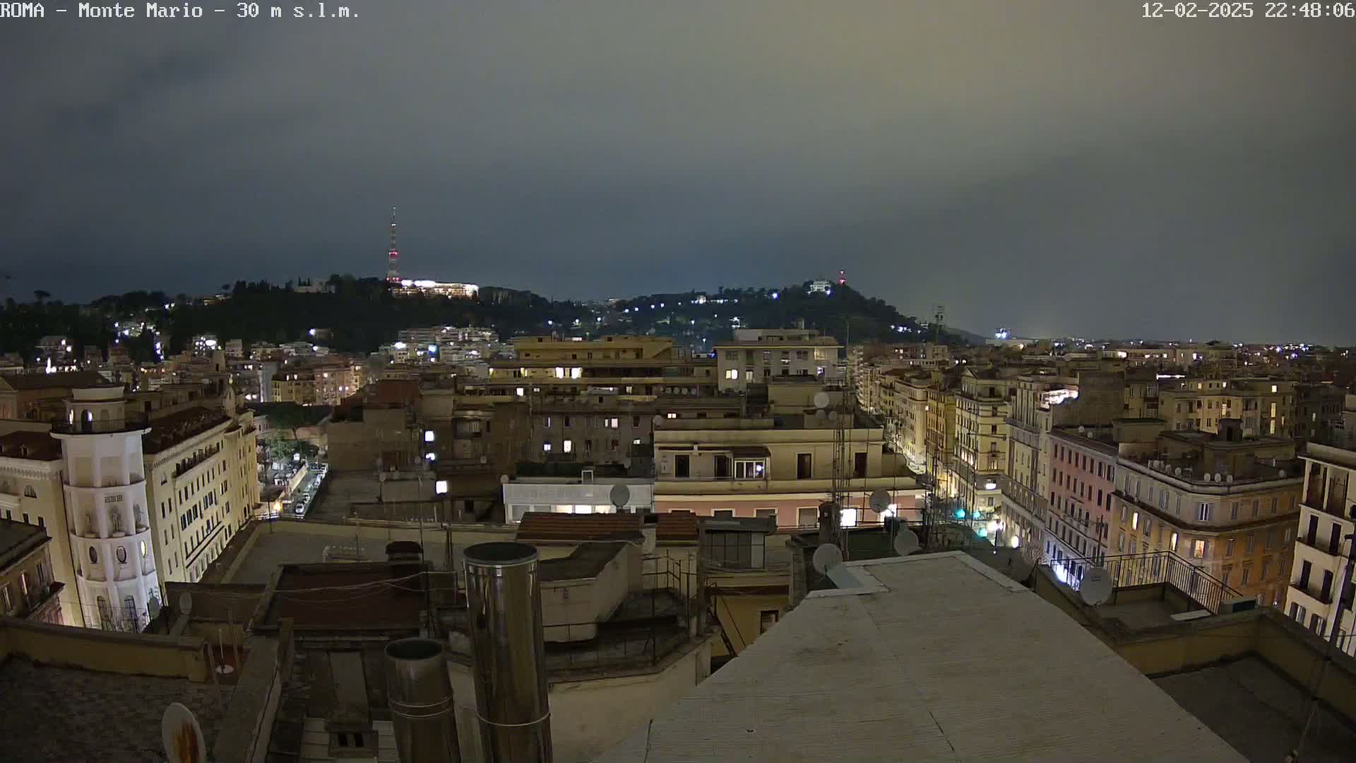 A nighttime view of a city, under an overcast sky, shows many buildings lit up with lights.