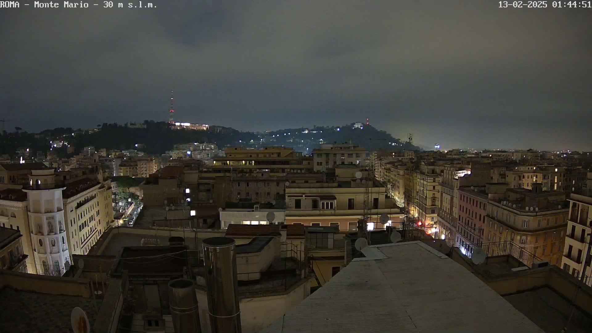 A nighttime view of a city with a hill in the background under a dark, cloudy sky.